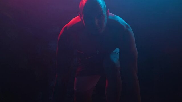 Male bodybuilder crouched down and getting ready to start, looking at the camera, in neon light in smoke in a dark room.