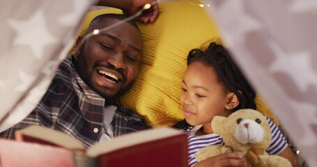 Happy african american father and daughter lying in tent and reading book - Powered by Adobe