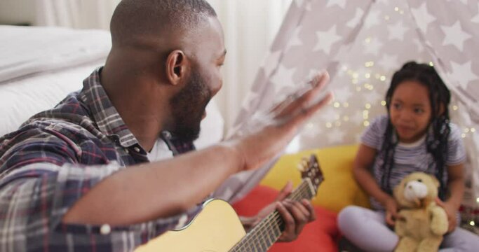 Happy african american father and daughter sitting in blanket tent, playing guitar, clapping hands