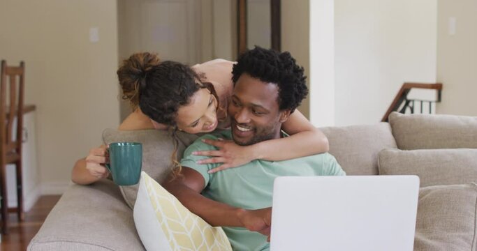 Happy biracial woman with coffee embracing her male partner sitting on sofa with laptop