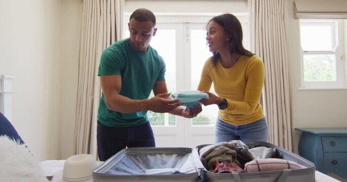 Happy biracial couple in bedroom packing clothes to suitcase for travel