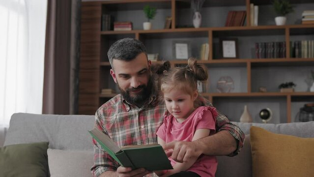 Young father and daughter read intersting book at home. Loving parent bearded man teaches his preschool child to read. Daddy reading fairy tale story. Parenthood, love and happy family concept.