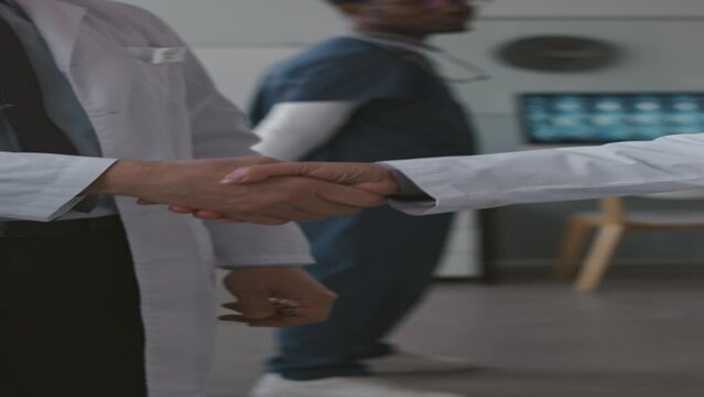 Vertical cropped slowmo of two unrecognizable medical colleagues wearing white lab coats shaking hands in office