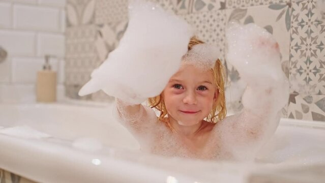 Cute caucasian blonde three years old girl taking bath playing with white soap foam bubbles,looking at camera.Carefree childhood, bathroom games concept.