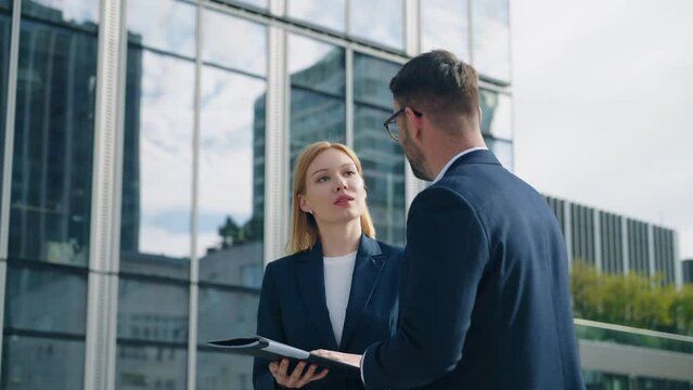 Gimbal shot two business partners caucasian man woman in formal suits standing outdoors office building terrace in downtown city discussing upcoming project and deals looking at paper documents folder