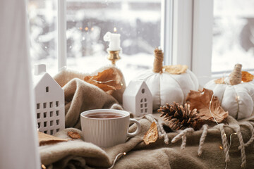 Fototapeta na wymiar Autumn still life. Warm cup of tea, cozy pumpkin pillows, fall leaves, candle, lights and cute buildings decoration on brown scarf on windowsill. Hygge fall home decor. Happy Thanksgiving