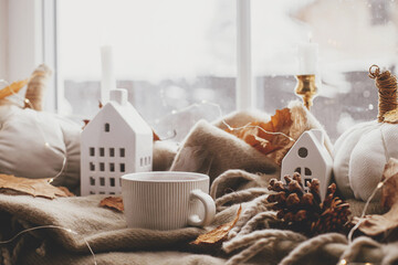 Fototapeta na wymiar Autumn still life. Warm cup of tea, cozy pumpkin pillows, fall leaves, candle, lights and cute buildings decoration on brown scarf on windowsill. Hygge fall home decor. Happy Thanksgiving