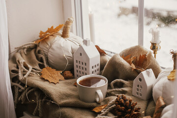 Cozy Autumn. Warm cup of tea, stylish pumpkin pillows, fall leaves, candle, lights and cute buildings decoration on brown scarf on windowsill. Autumn hygge, fall home decor