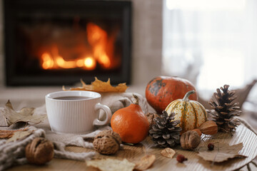 Cozy autumn. Warm cup of tea, pumpkins, autumn leaves, cones, cozy scarf on rustic wooden table on background of fireplace in farmhouse. Comfort fall in rural home. Happy Thanksgiving