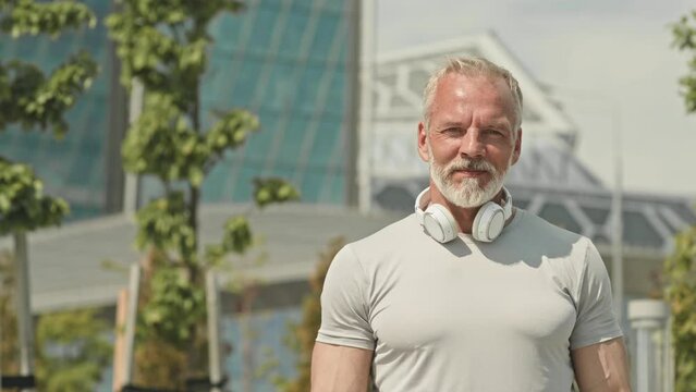 Portrait of senior athletic man taking off wireless headphones and posing for camera with smile while standing outdoors on city streetPortrait of senior athletic man taking off wireless headphones and