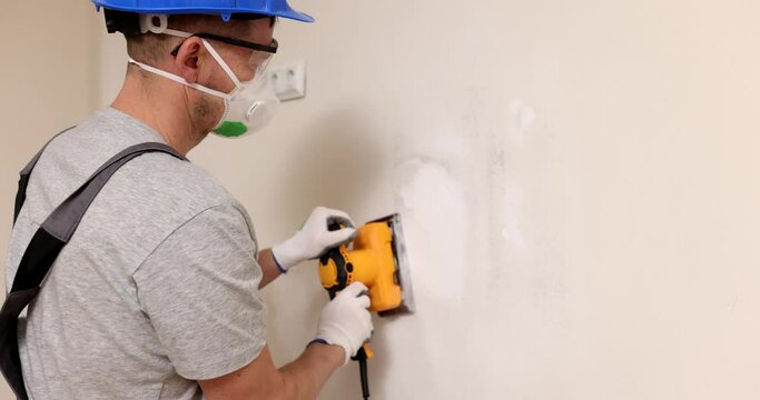 Labourer in helmet with respirator polishes plaster coverage on wall in room. Builder fixes holes on drywall panel with electrical grinder tool