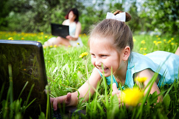 Young girl with laptop resting on a green lawn or a field in a park in nature and her mon on the background. Fmily with mother and daughter chatting, working, communicating in Internet