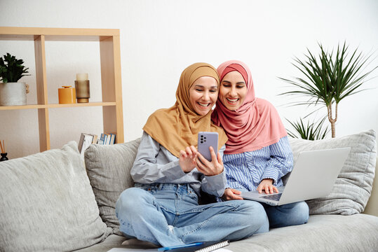 Portrait of beautiful happy arab girls in hijab working at home using smartphone and laptop while sitting on sofa. Women looking and typing on her mobile phone, holds a laptop