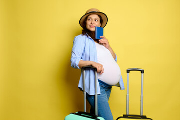 Delightful pregnant woman traveler smiles, dreamily looks aside a copy space, posing with suitcases on yellow background