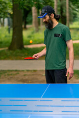 An elderly man next to a blue ping pong table hits an orange ball on a tennis racket in a city park