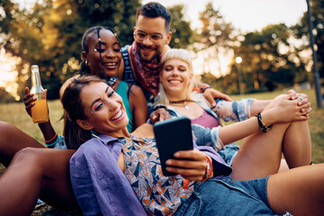Fototapeta premium Happy woman taking selfie with her friends while relaxing on grass during summer music festival.
