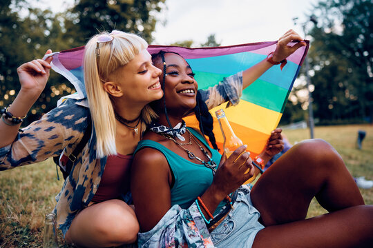 Happy lesbian couple with rainbow flag relaxing on grass during summer music festival.