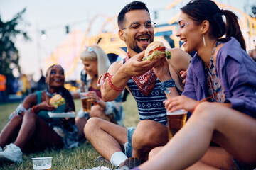 Group of happy festival goers eat hamburgers while relaxing on grass.
