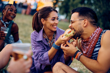 Fototapeta na wymiar Happy couple sharing hamburger while attending summer music festival with their friends.