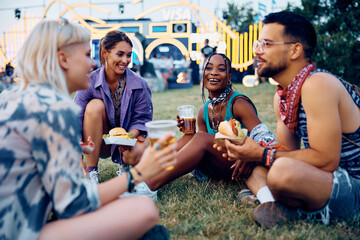 Multiracial group of happy festival goers eat burgers and drink beer while relaxing on grass.