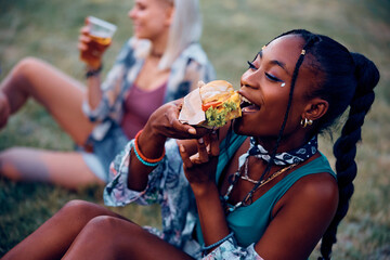 Happy black woman eats hamburger while attending summer music festival.