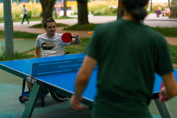 Inclusiveness Happy A disabled man in a wheelchair plays ping pong against an old man with a gray beard in a city park 