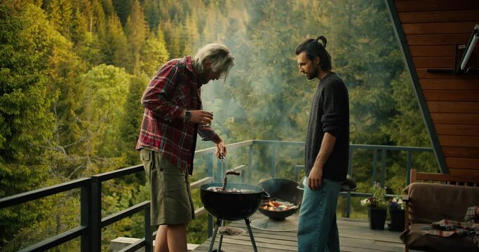Two guys fry sausages on the grill on the grill. A man in a red shirt turns over sausages using special tongs against the backdrop of mountains and forest