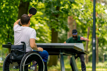 Inclusiveness A disabled man in a wheelchair plays ping pong with an older man in a city park against a backdrop of trees