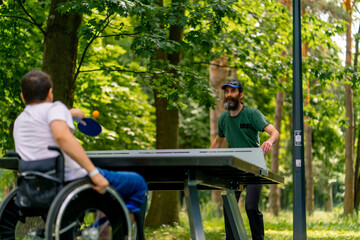 Fototapeta premium Inclusiveness A disabled man in a wheelchair plays ping pong with an older man in a city park against a backdrop of trees