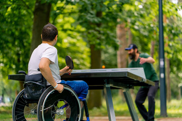 Inclusiveness A disabled man in a wheelchair plays ping pong with an older man in a city park against a backdrop of trees