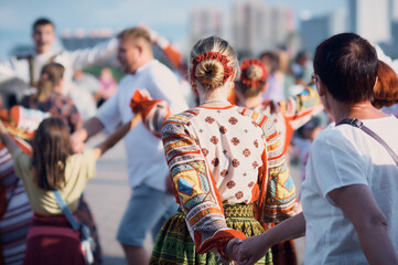 Folklore ensemble of a round dance on the embankment. Spectators dance along with dancers in national costumes. Bright sunlight. Concept: Summer holidays and festivals around the world. Holding hands