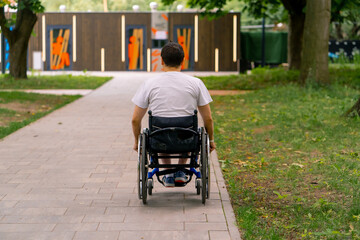 Obraz premium Inclusiveness A man with disabilities rides a wheelchair along the path of a city park against the background of trees Back view