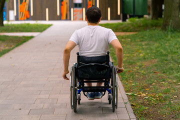 Obraz premium Inclusiveness A man with disabilities rides a wheelchair along the path of a city park against the background of trees Back view