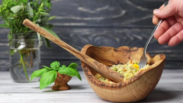 A man taking a forkful of vegetarian couscous from a wooden bowl, with a dark wood background and herbs.
