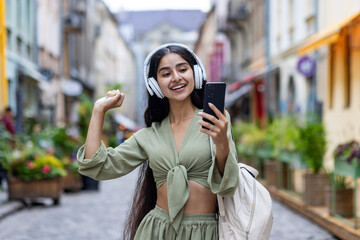 Young beautiful id woman walks in the city, tourist holds phone in hands, uses headphones to listen...