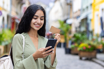 Young smiling Indian woman walking in the city, woman holding a bank credit card and phone, tourist...