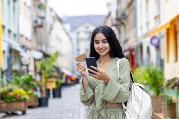 Young smiling Indian woman walking in the city, woman holding a bank credit card and phone, tourist making online booking of accommodation and booking tourist services while walking in the city - obrazy, fototapety, plakaty