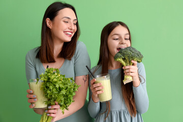 Little girl with her mother and vegetables drinking smoothie on green background