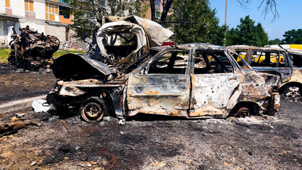 Fototapeta premium Burned-out cars at the site of the missile strike. In the background are residential buildings with broken windows. Russian-Ukrainian War 2022-2023. Missile attack on residential buildings in