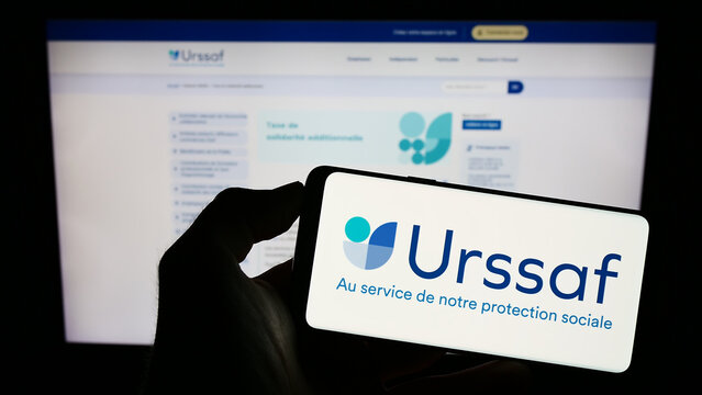 Stuttgart, Germany - 07-12-2023: Person holding cellphone with logo of French social security organization URSSAF on screen in front of webpage. Focus on phone display.