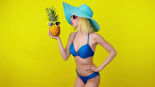 Sexy woman in bikini dancing fashionable hat with pineapple, smiling enjoys summertime for spa and wellness. Playful glamor lady dance, slim body for tan or pool party, colorful trend on vivid yellow