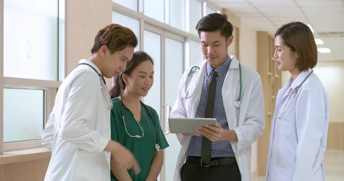 A group of Asian male and female doctors standing talking and giving opinions about treating patients with a smile in hospital hallway. Medical Team using digital tablet and discussing.