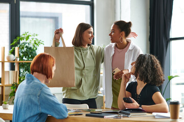 excited woman showing shopping bag and smiling at african american girlfriend near multiethnic women sitting at table in friendly atmosphere of interest club, sharing joy and positive emotions