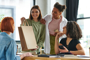 young and cheerful woman standing with shopping bag near african american girlfriend with smartphone and multiethnic women spending time in interest club, sharing joy and positive emotions