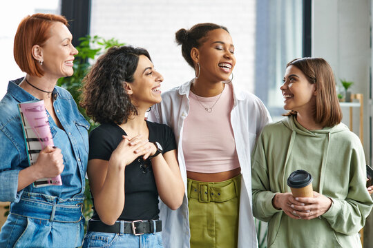 young and happy woman with coffee to go looking at stylish multiethnic female friends standing and laughing in women interest club, mutual support, solidarity and trust concept