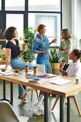 cheerful redhead woman with smartphone talking to laughing friend near multicultural women drinking coffee from paper cups in women interest club, happy gathering and leisure of diverse female team