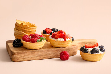 Wooden board of tasty tartlets with whipped cream and berries on beige background