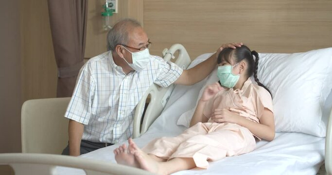 Asian senior older grandfather wearing a face mask and glasses talking, playing, taking care and comforting a little sick girl in patient room at hospital. Healthcare and emotional expression concept.