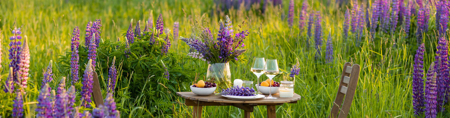 Beautiful romantic outdoor wedding decor in field. Table decorated with purple lupines flowers. Wineglasses with white wine. Sunset, summer, golden hour. Perfect surprise date for loving couple banner