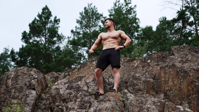 Caucasian strongman stands on the high rock. Sportsman with naked torso demonstrating his athletic body. Low angle view.